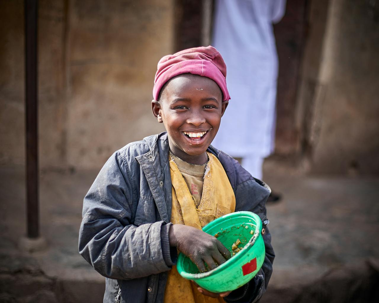 Happy Nigerian child holding a bowl outdoors in Kaduna, wearing a colorful beret and jacket.
